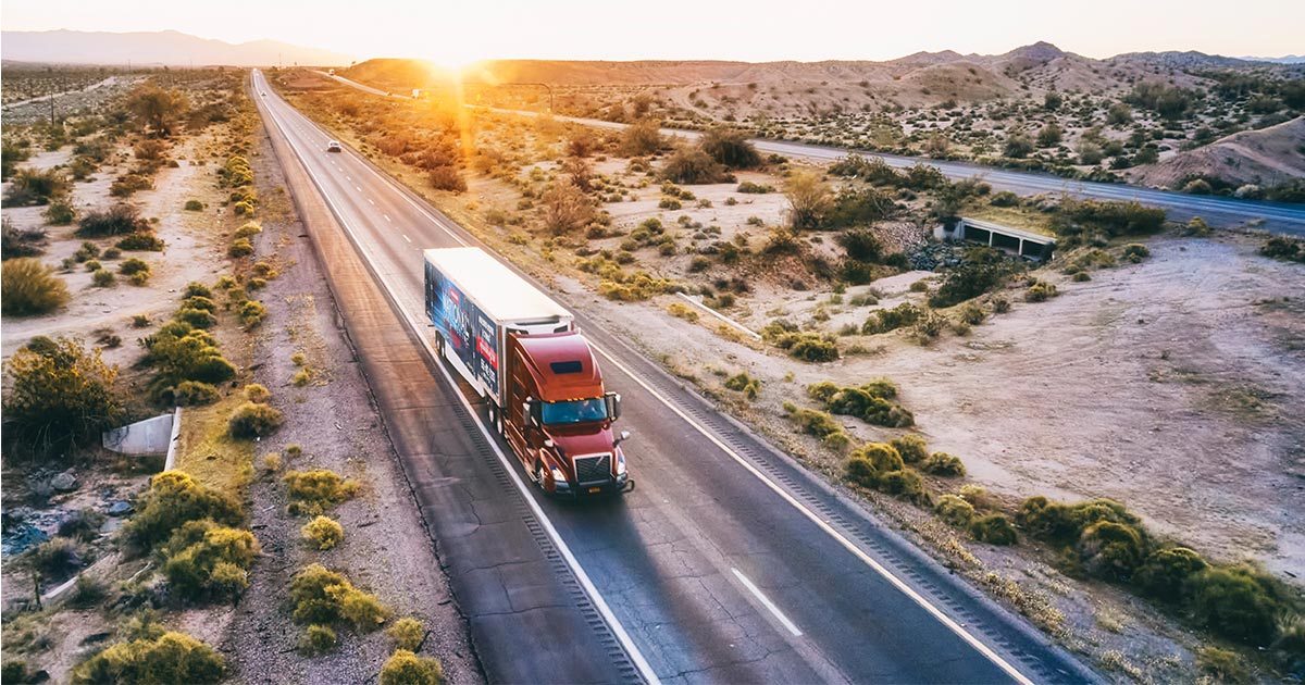 A National Van Lines moving truck drives down a desert highway. The sun is setting behind the truck on the horizon. 
