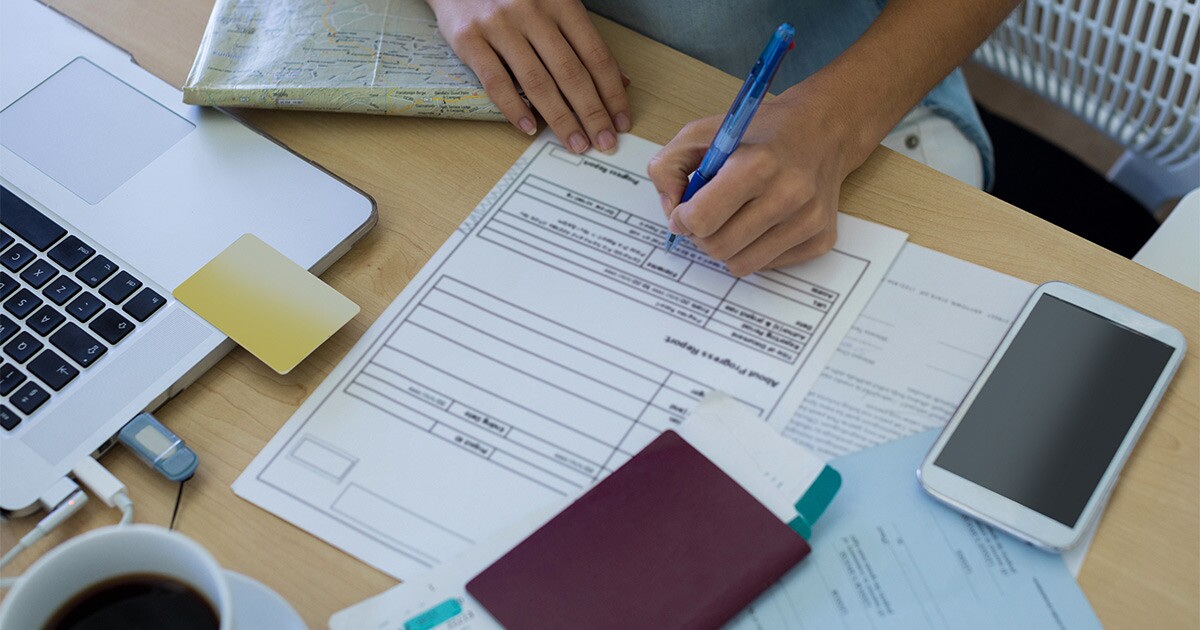 Someone sits at a desk with their laptop. They have multiple documents in front of them, including paperwork, a passport, and plane tickets. They are preparing to move out of the country, and are calculating international moving costs.