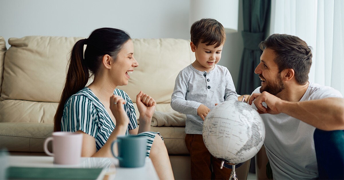 Two smiling parents sit with their young child in front of a couch in a living room. They are showing the child a globe and discussing their plans to move out of the country. Because they have properly calculated international moving costs, they are now getting excited about their move to a new country.