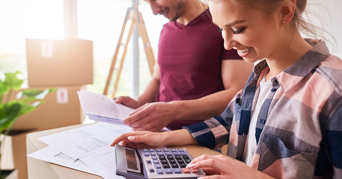 A smiling couple sits on a couch amongst moving boxes. They are reviewing paperwork and calculating their international moving costs for their upcoming move out of country.