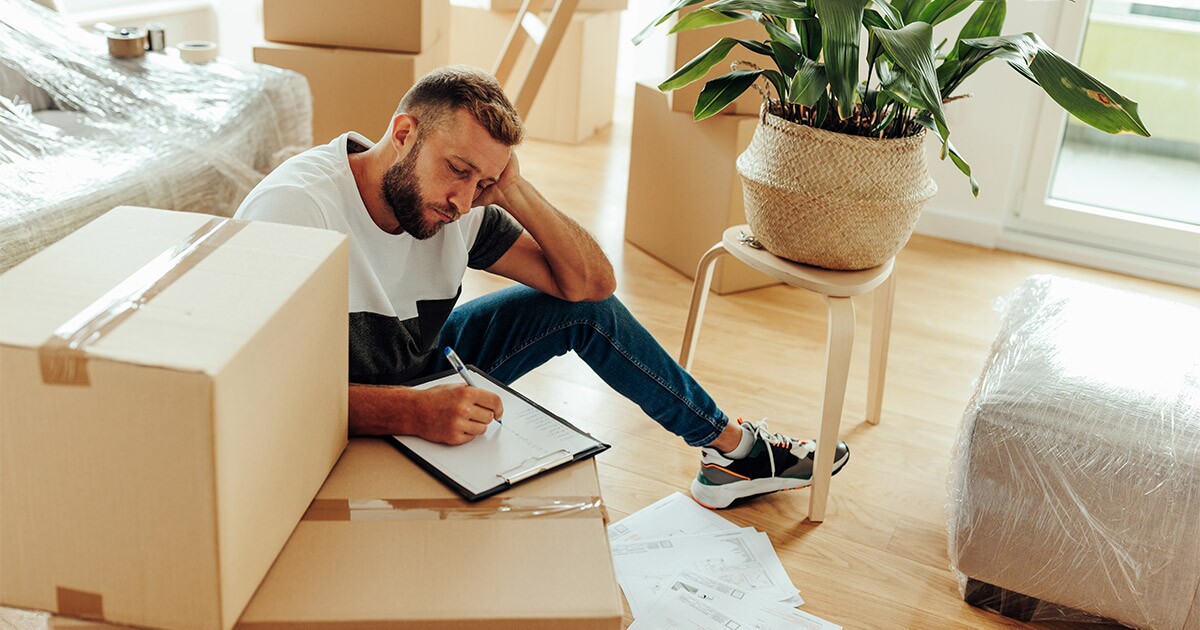 A man sits amongst moving boxes and packing supplies, and writes on a clipboard. The clipboard lists what movers won't move. He is preparing for an upcoming long distance move.