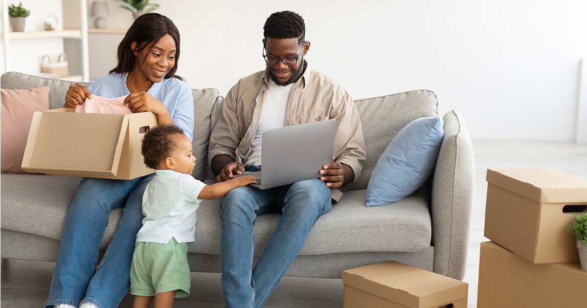 Two parents sit on a couch. The mother folds clothes into a moving box while the father shows their baby something on his laptop computer. They are planning for a long distance move.