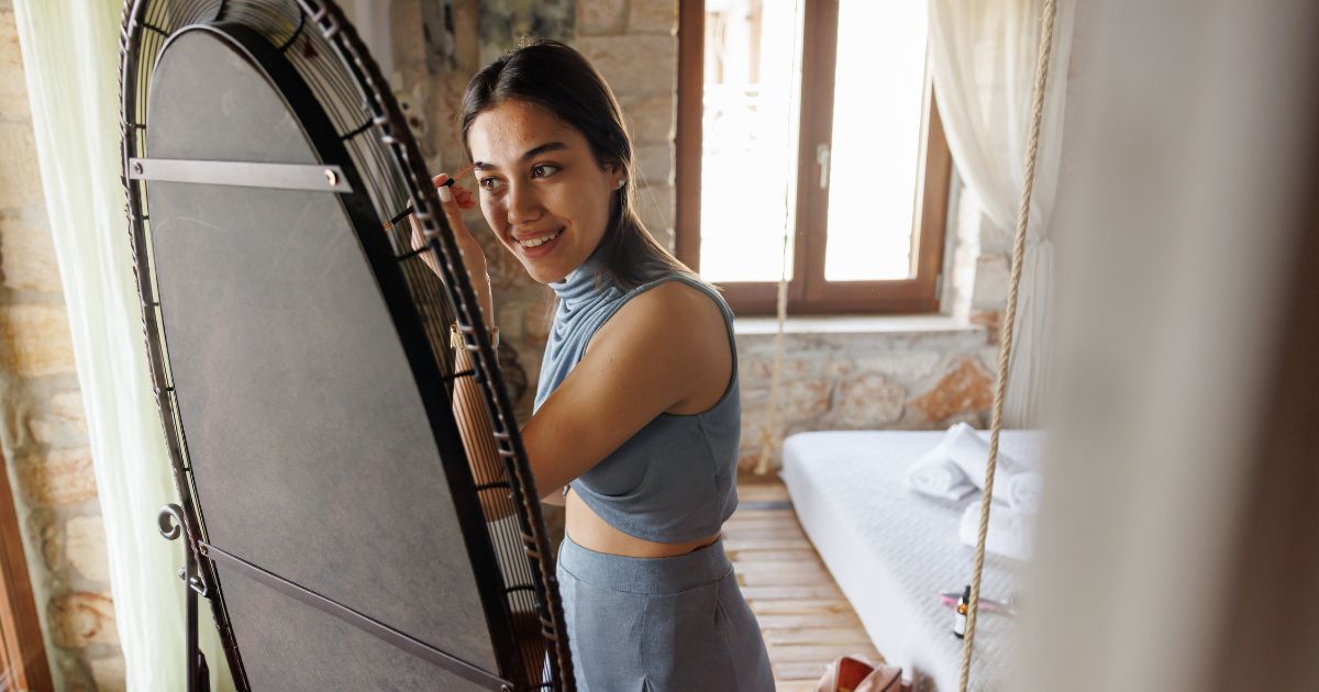 A young woman, who has just completed a long distance move, puts on makeup in her full length mirror. She is smiling because moving her mirror went well.
