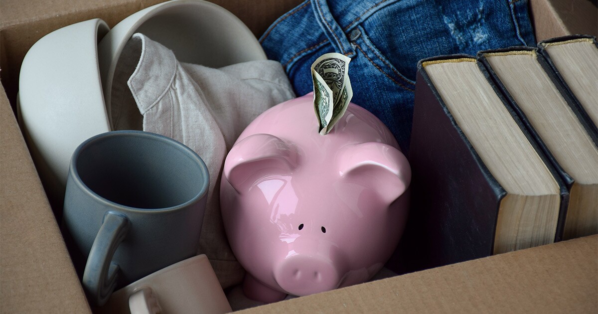 A piggy bank sits in a moving box amongst items like books, cups, and clothing. A dollar bill is sticking out of the top of the piggy bank. This will be used as part of the tip someone leaves for a moving crew after a long distance move.
