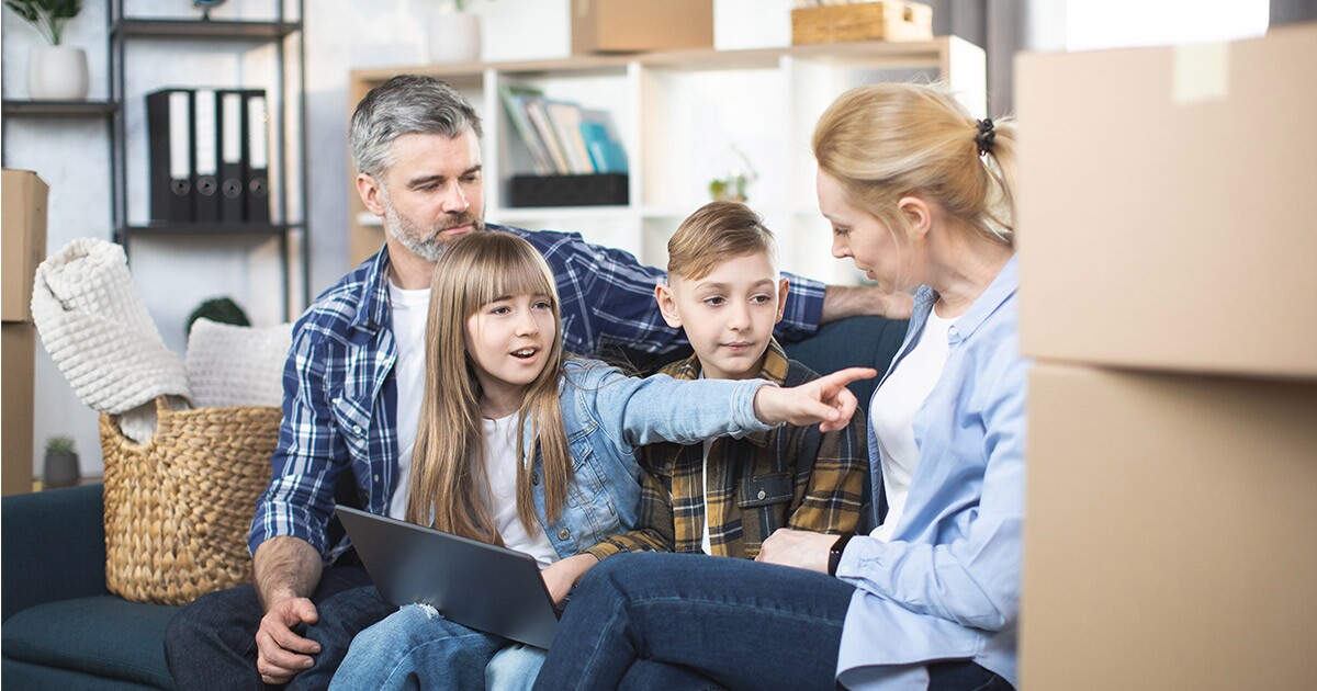 A family of four sits on their couch. The daughter has a tablet on her lap and is pointing towards some moving boxes next to her mother. The family is discussing moving cross country, and are figuring out exactly where they want to start planning. 