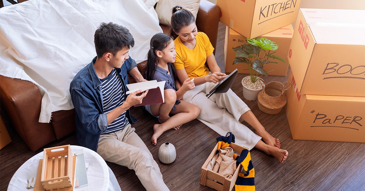A family of three sits amongst moving boxes and packing supplies. They are getting ready to book their long distance move that is occurring in a few months. 