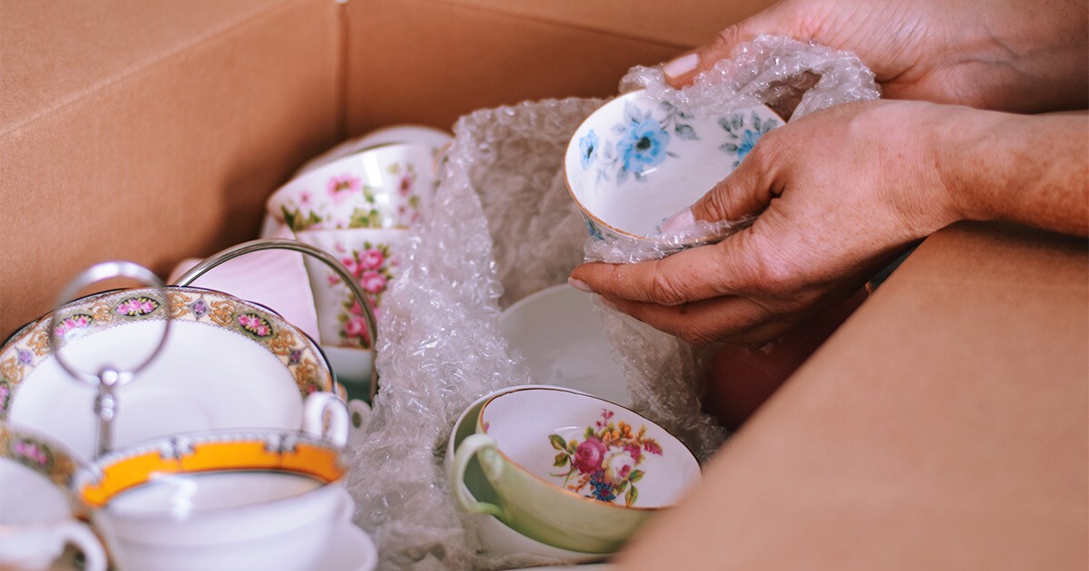 Someone carefully places bubble-wrapped porcelain dishes into a moving box for a cross country move. They are moving antiques as a part of this.